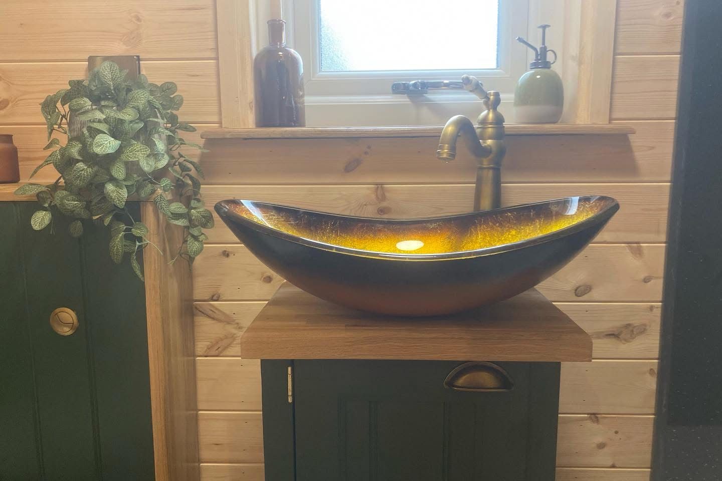 Rustic bathroom with a copper vessel sink, brass faucet, wood walls, and potted plants on the counter.