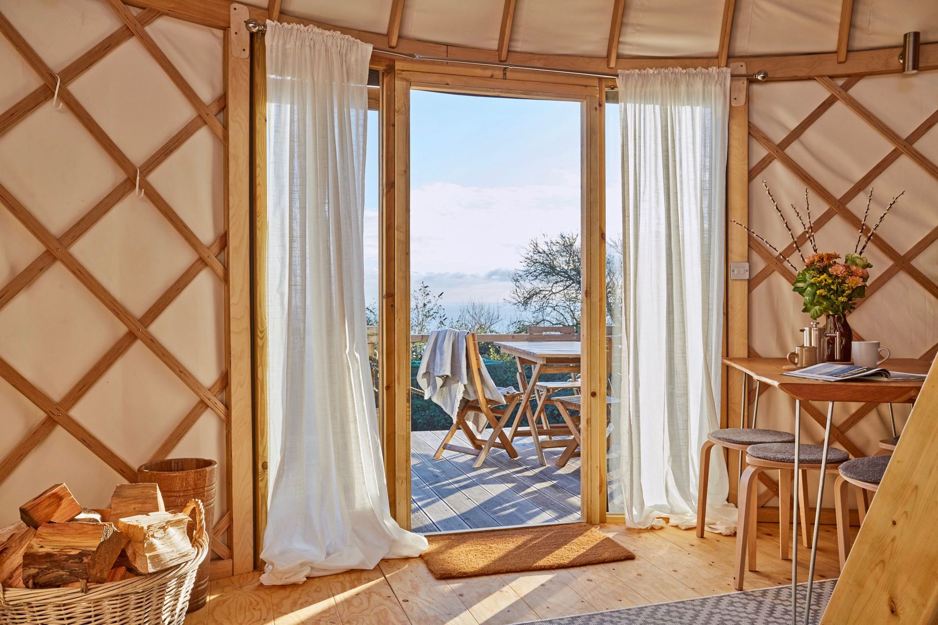 Cozy yurt interior with open doorway to a sunlit deck, sea view, and white curtains.