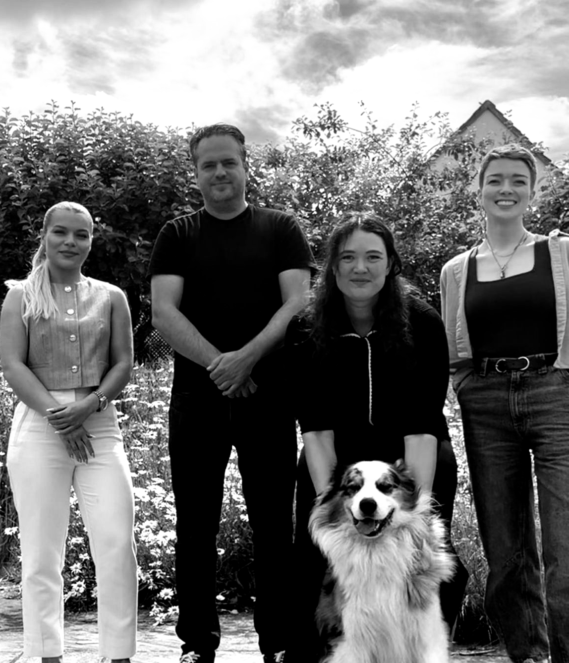 Four people and a dog posing outdoors in front of trees and a house