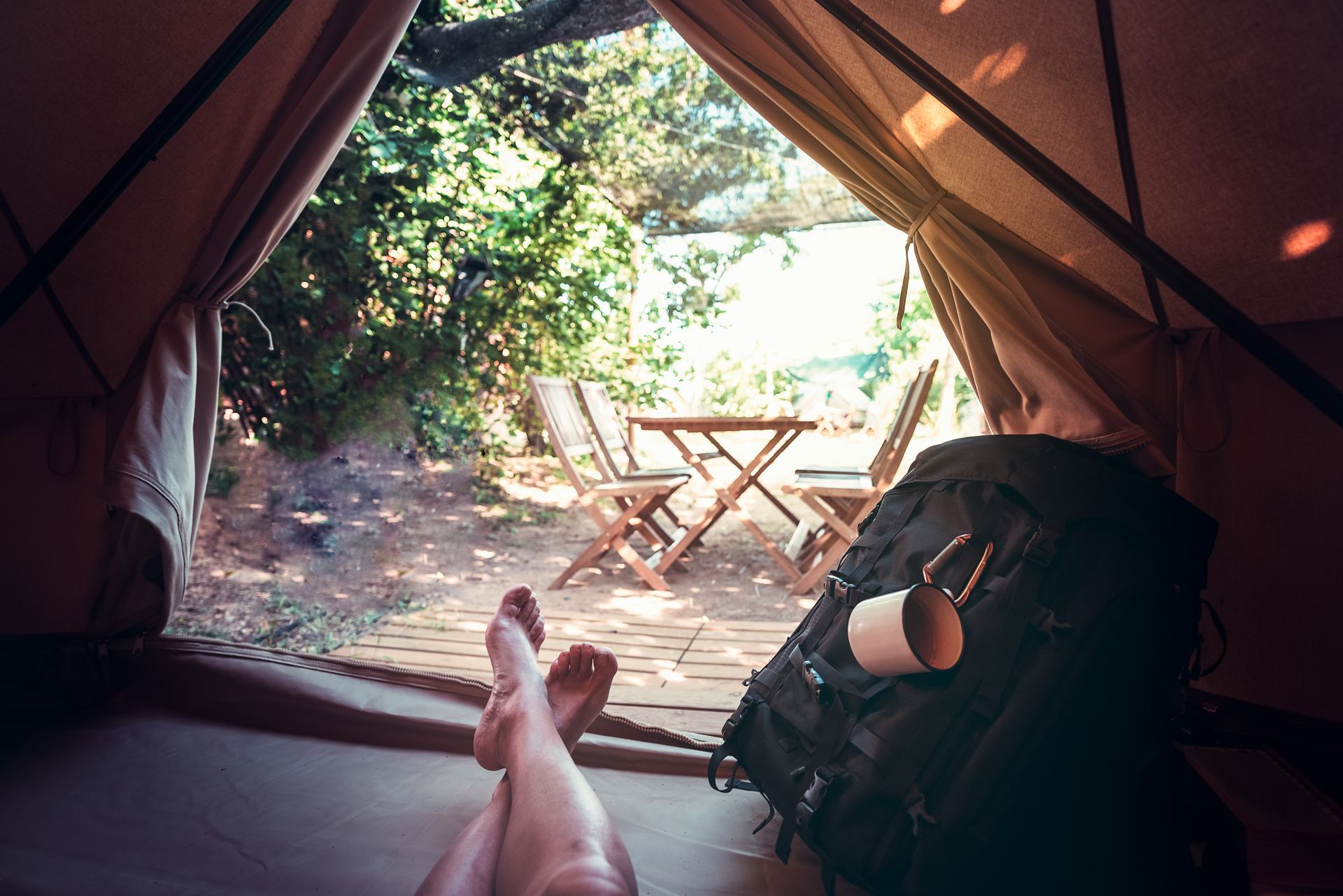 Inside a tent, a person’s feet rest toward a wooden table and chairs in a sunny forest campsite.