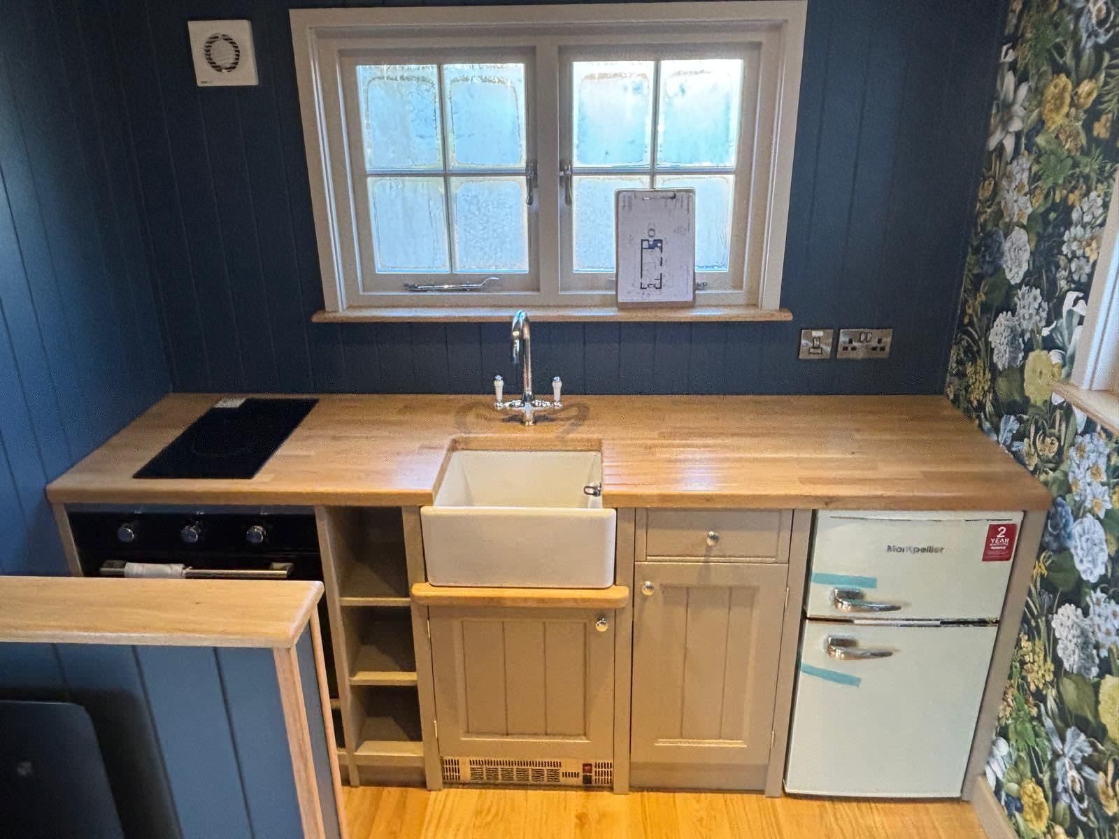 Small kitchen with wooden counters, white sink under a window, black stove, and floral wallpaper.