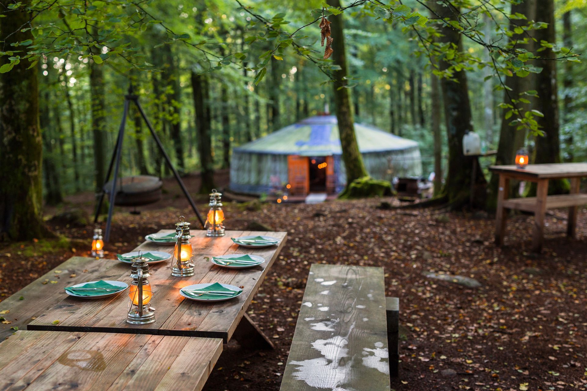 Woodland campsite with a lit yurt and long wooden tables set with lanterns and plates