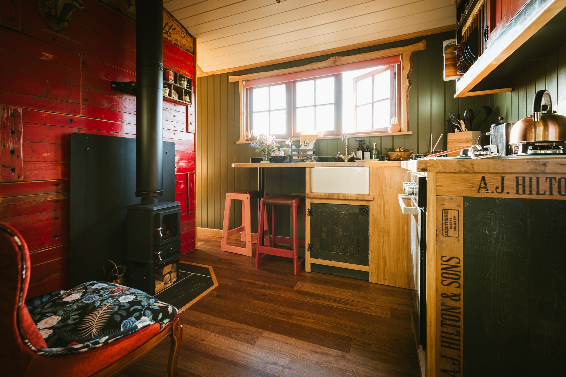 Cozy rustic kitchen with red walls, wood stove, sunlit window, and wooden counter and stools