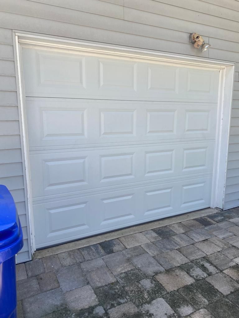 A white garage door is sitting on the side of a house next to a blue trash can.