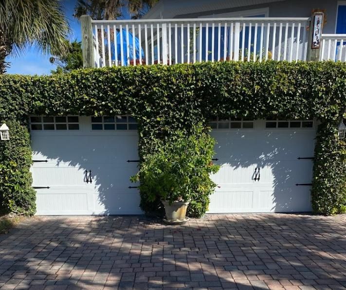 A white garage door is surrounded by ivy and a brick driveway