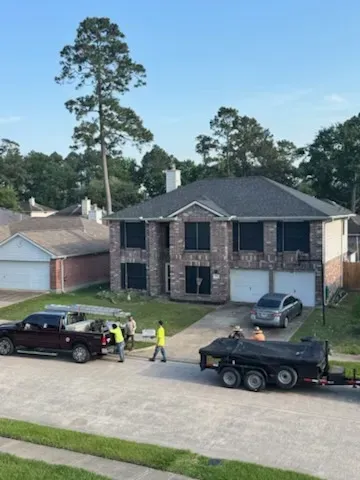 Two-story brick house with workers, a truck, and a trailer in front. Suburban setting, sunny day.