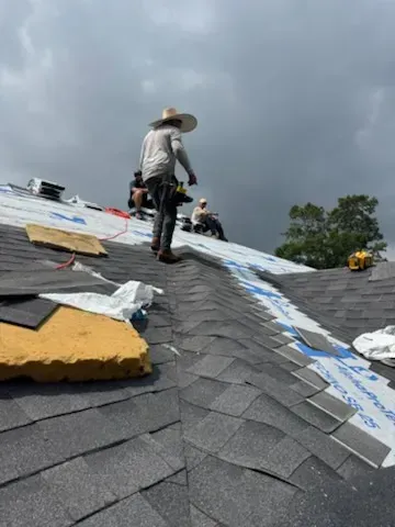 Roofers working on a dark gray shingled roof under a cloudy sky.