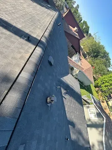 View of a dark shingled roof, vent pipes, and another house against a hillside with trees.