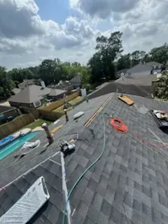 Rooftop with gray shingles, tools, and hoses. Houses and trees visible in the background under a cloudy sky.