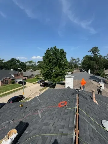 Roofers working on a dark shingle roof on a sunny day. Houses and trees in the background.