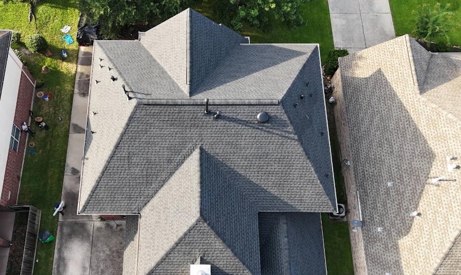 Overhead view of a dark gray shingled roof with vents on a suburban home. Green grass and neighboring houses are visible.