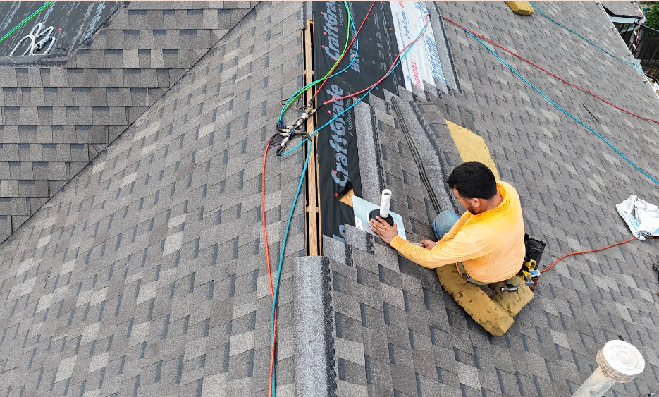A roofer is installing gray shingles on a roof ridge. They are using safety lines and a wooden platform.