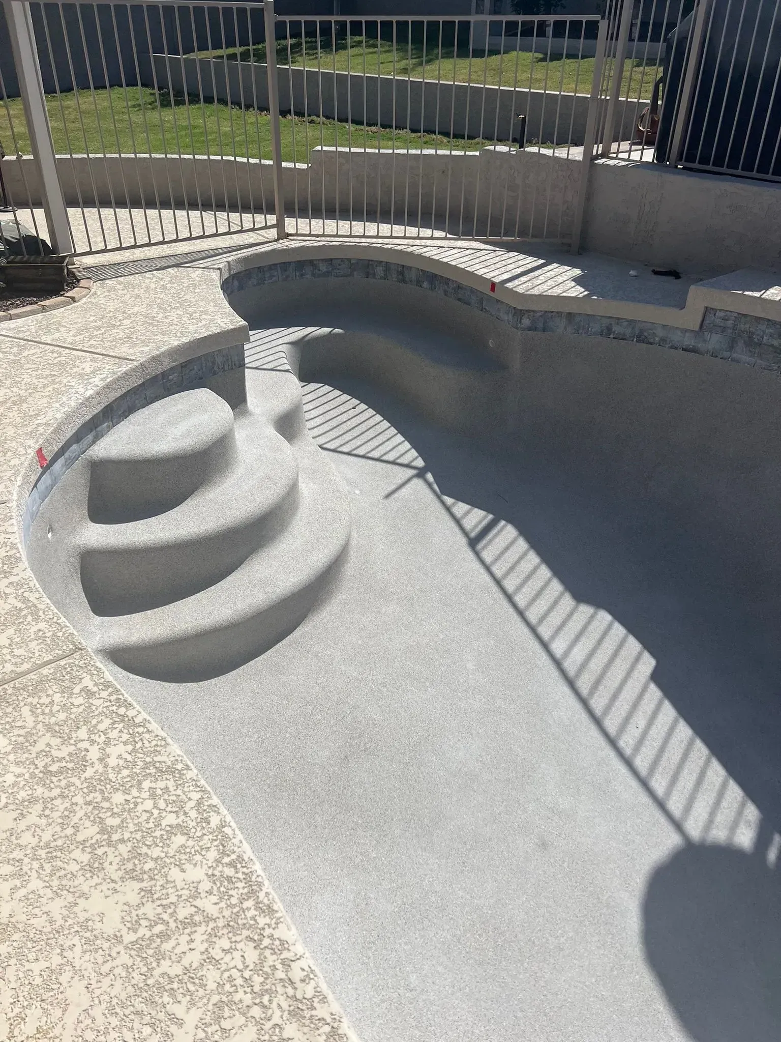 Empty, gray swimming pool with steps, surrounded by light-colored textured concrete and a metal fence.