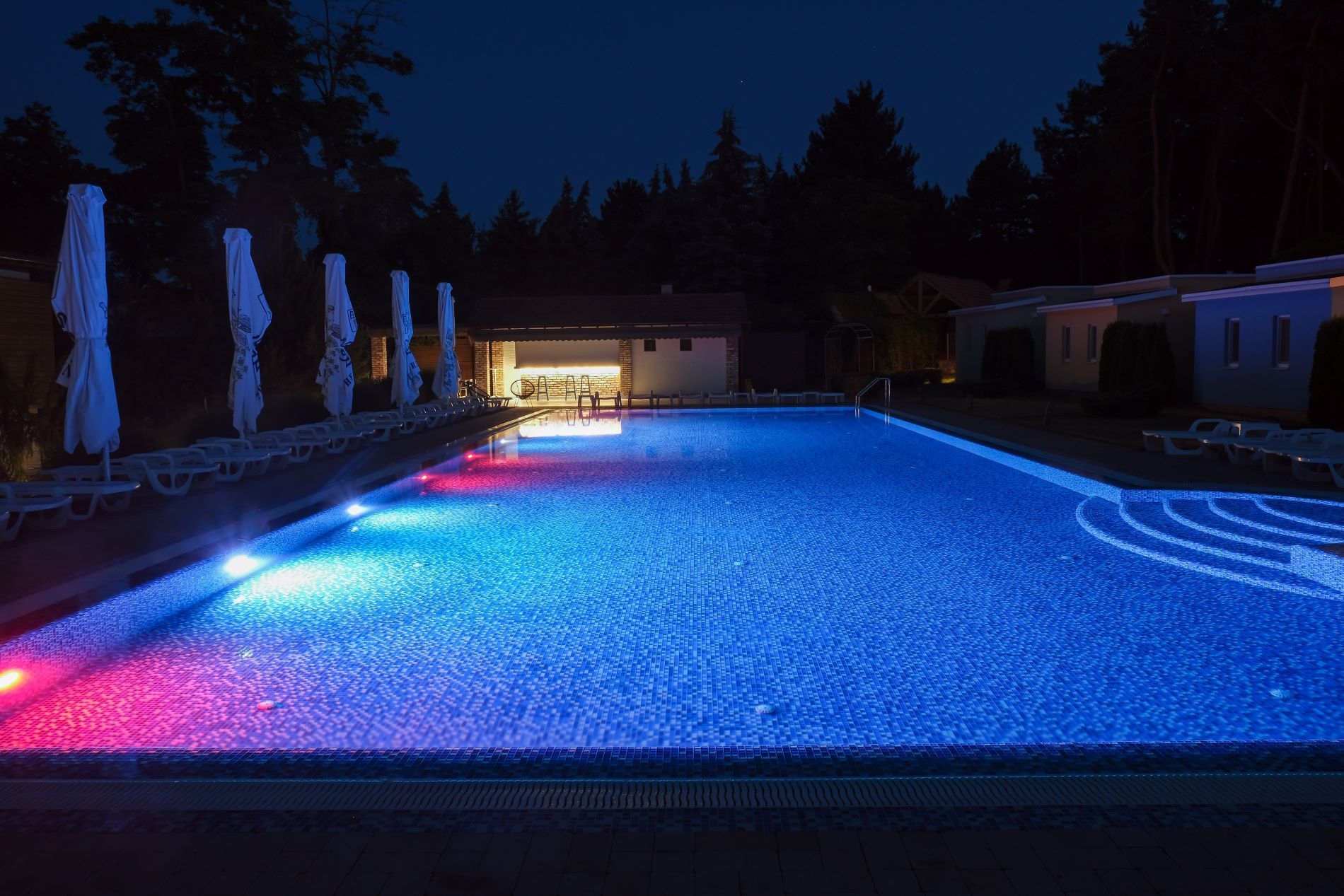 Nighttime illuminated swimming pool with red and blue lights, next to a bar and cabins.