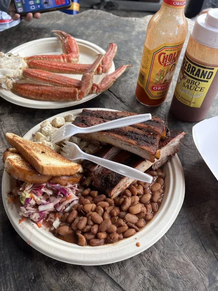 Ribs, baked beans, coleslaw, and crab legs on paper plates with hot sauce bottles.