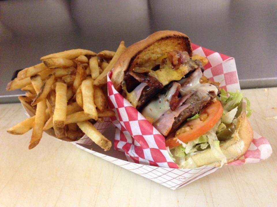 Burger with fries in a checkered paper basket. The burger has multiple layers of meat, cheese, lettuce, tomato and bacon.