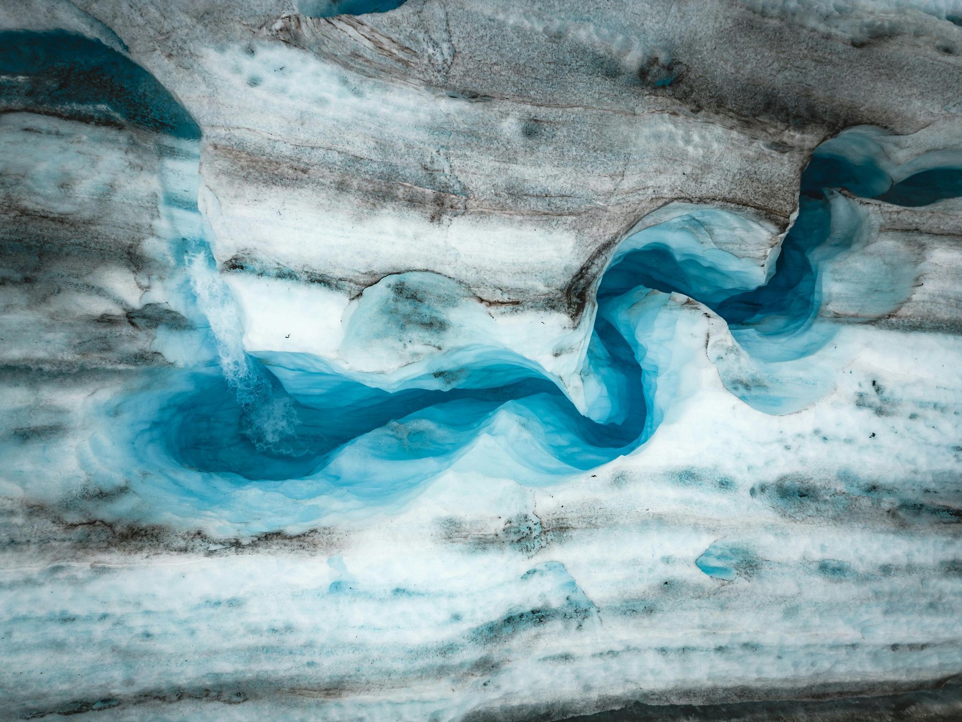 A blue glacial stream flowing across a textured icy landscape, with shades of white, gray, and blue.