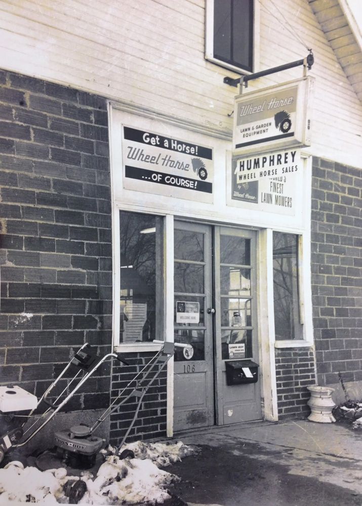 Exterior of a brick building with signs for car repair and a sign swinging above the entrance. Two lawnmowers are visible.
