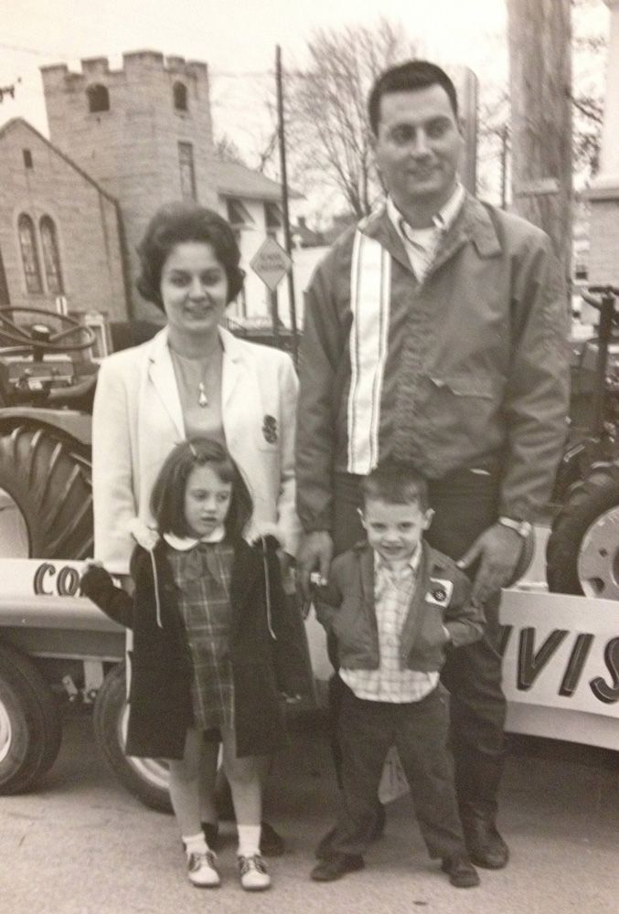 Family of four poses near a tractor. Mother in a suit, father in a jacket, and two children hold hands.