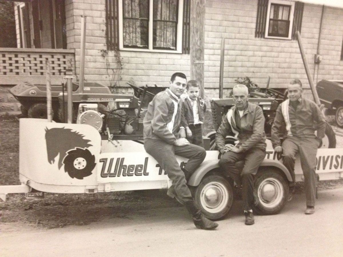 Four men pose on a trailer.  They are in front of a brick building.