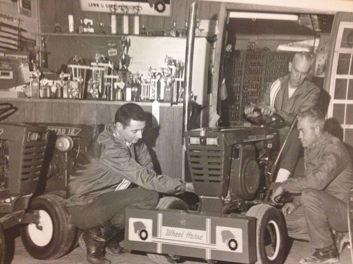 Three men working on a racing vehicle inside a garage. One man kneels, two stand, all focused on the project.