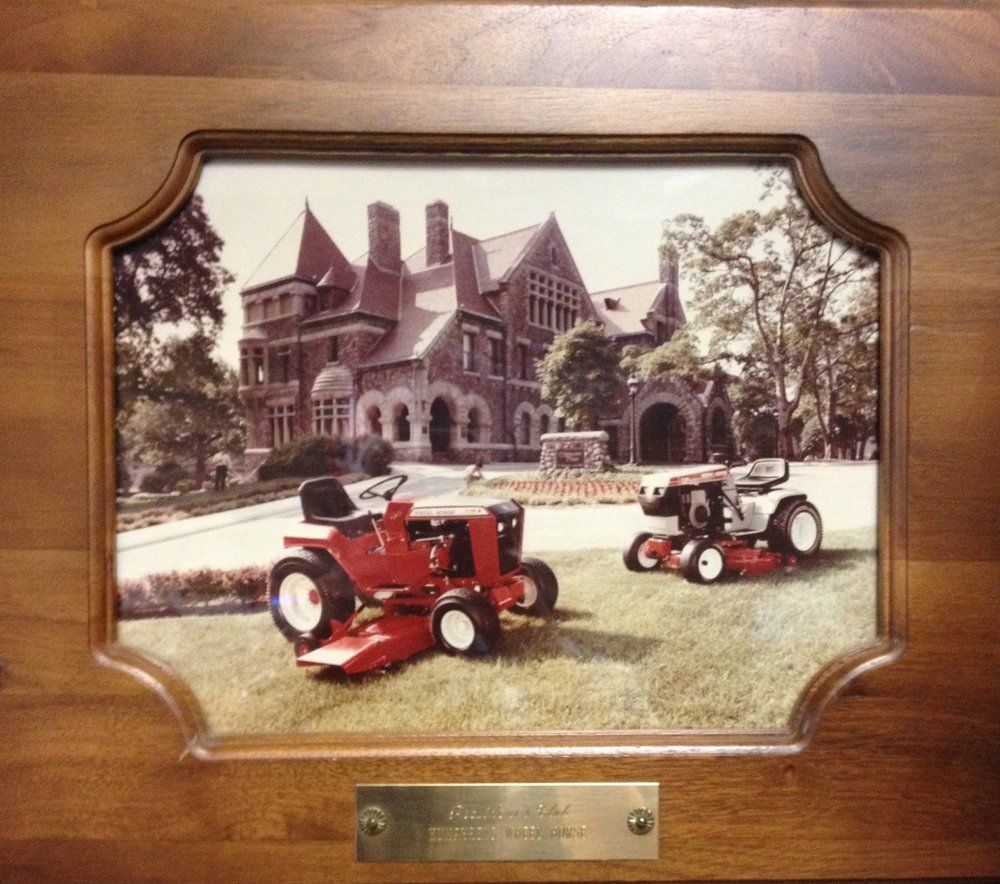 Two lawn tractors in front of a large mansion; framed photo in a wooden plaque.
