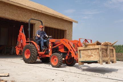 Orange tractor carrying a wooden crate near a building; a person is operating it.
