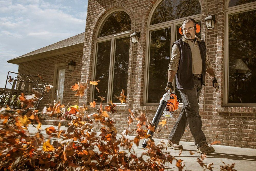 Man using a leaf blower on a brick patio, blowing fall leaves. Wearing ear protection, near a house.
