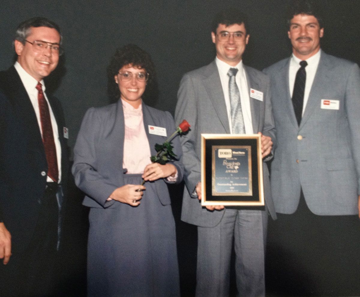 Four people, two men and two women, pose with an award. One woman holds a rose. They are standing indoors.