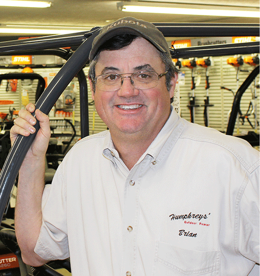 Man in a Kubota hat and button-down shirt smiles, holding a machine's handle in a store.