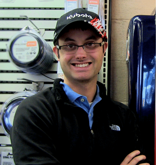 Smiling person in a Kubota hat and glasses, arms crossed, standing in a hardware store.