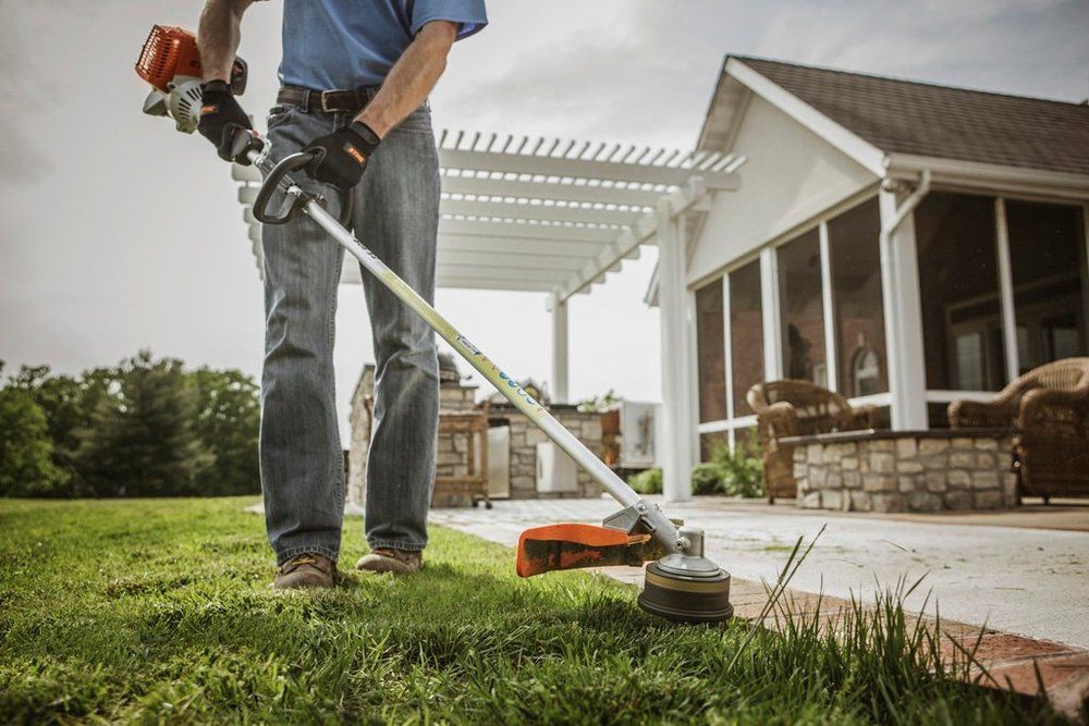 Person using a string trimmer to cut grass near a patio and house.