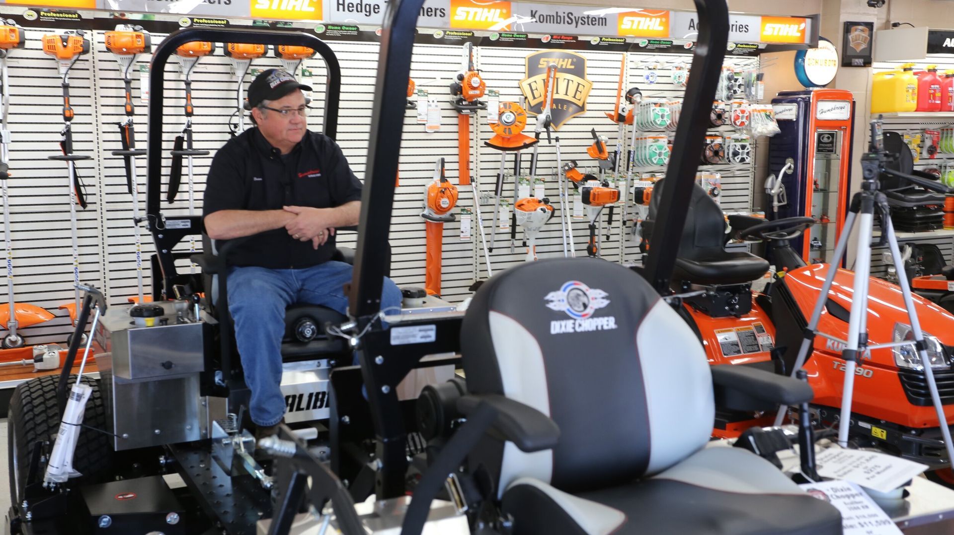 Man seated on a black zero-turn mower in a store; orange tools in background.