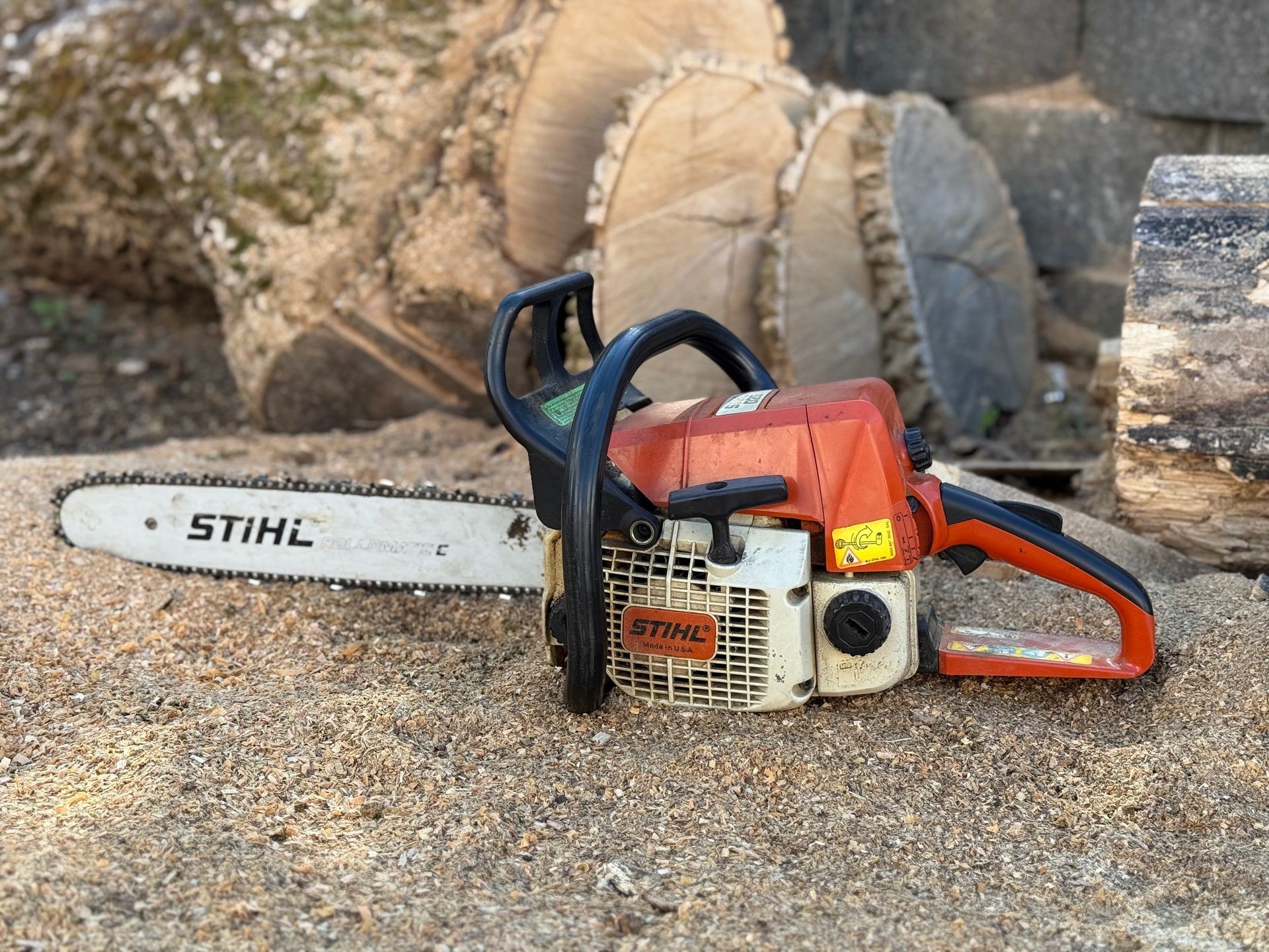 A Stihl chainsaw on wood chips, with a tree trunk in the background. Orange and white colors.