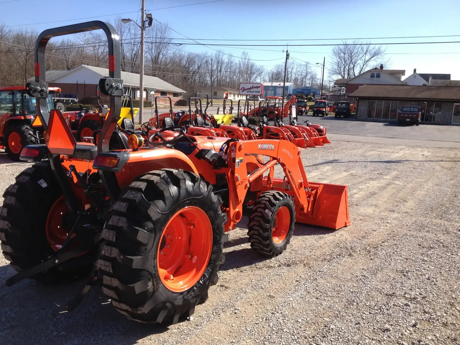 Orange tractor with bucket parked on a gravel lot; other tractors and a building are visible.