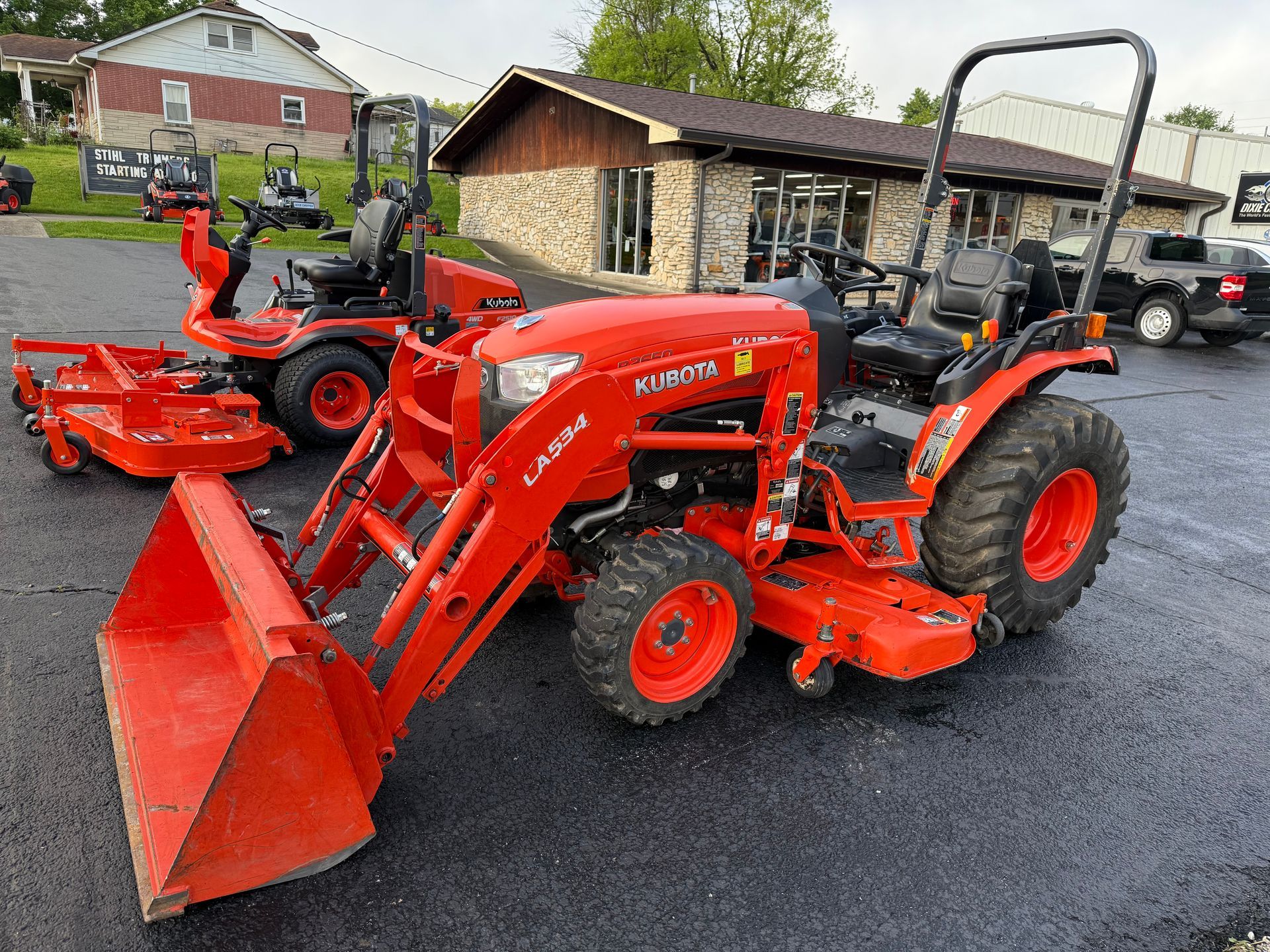 Orange tractor with front loader and backhoe parked in front of a building with a "Dixie Chopper" sign.