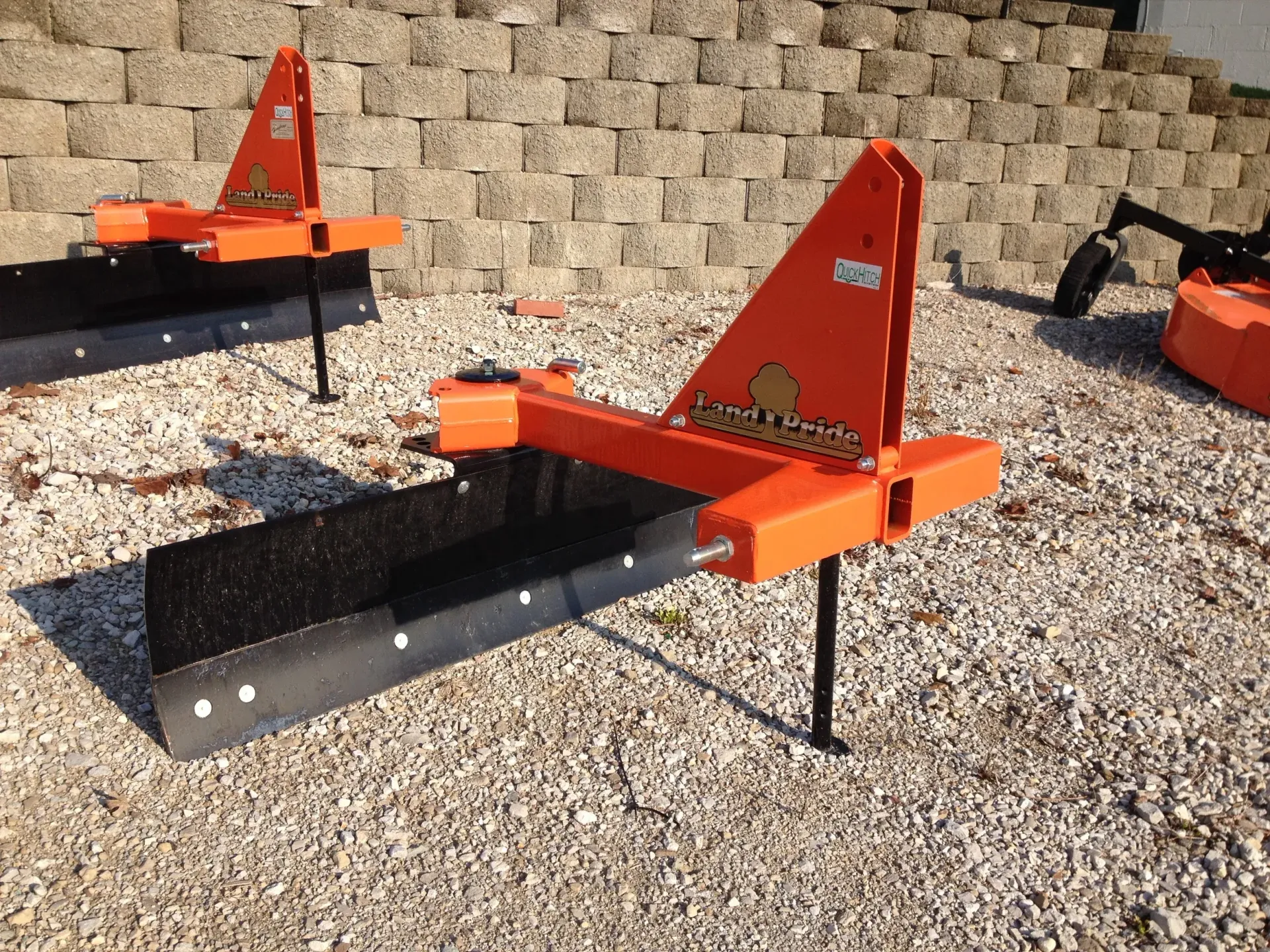 Orange and black land grading blades, angled against a stone wall background.