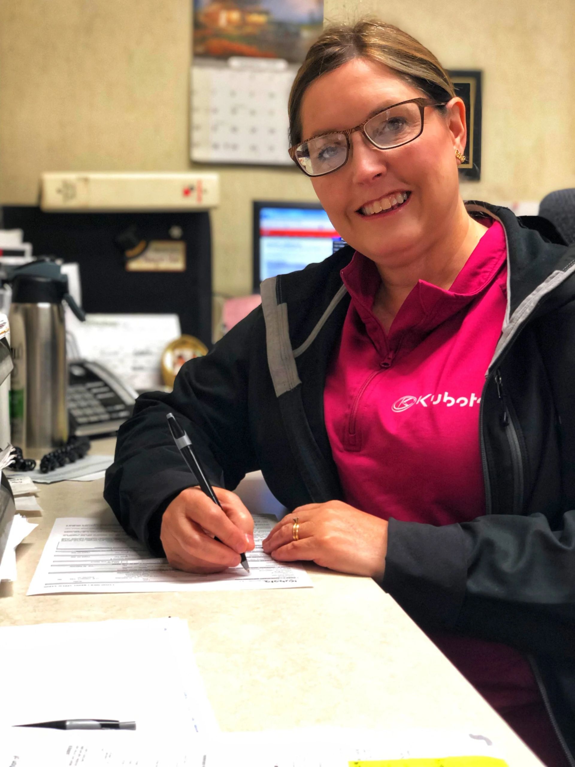 Woman with glasses, smiles, writing at a desk, wearing pink shirt and black jacket.