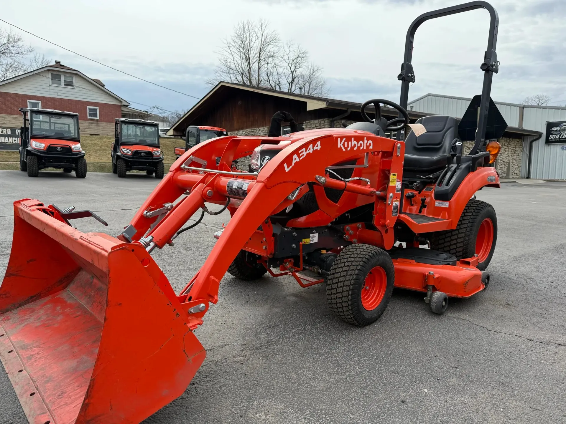 Orange Kubota tractor with front loader, parked on asphalt; two utility vehicles in the background.