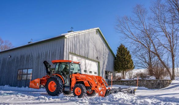 Orange tractor plowing snow in front of a metal building on a sunny winter day.
