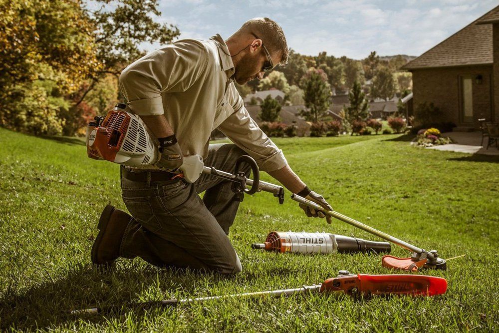 Man in a yard, kneeling, inspecting string trimmer and attachments.