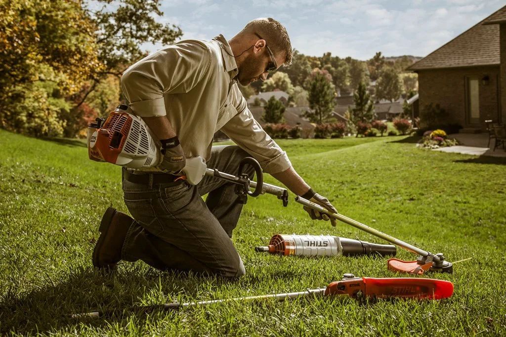 Man kneeling, assembling trimmer in grassy yard near house, bright day.