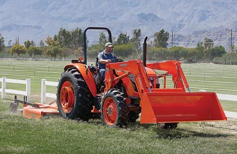 Orange tractor mowing grass in a field with a man driving. White fence and mountains in the background.