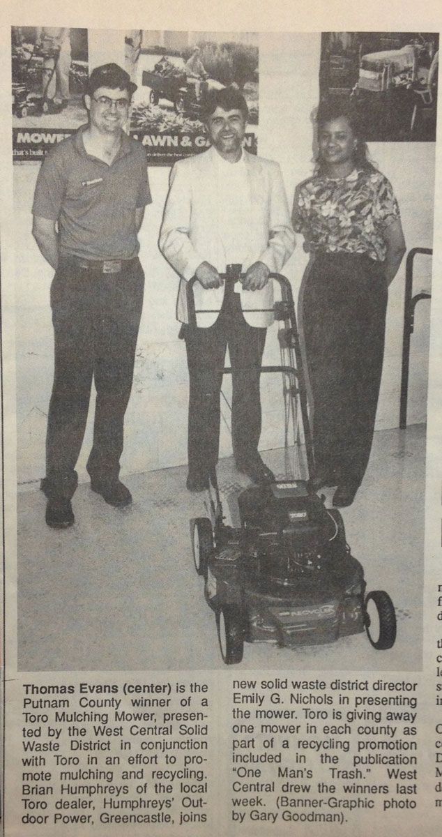 Three people standing with a lawnmower inside a building. Two men and one woman.