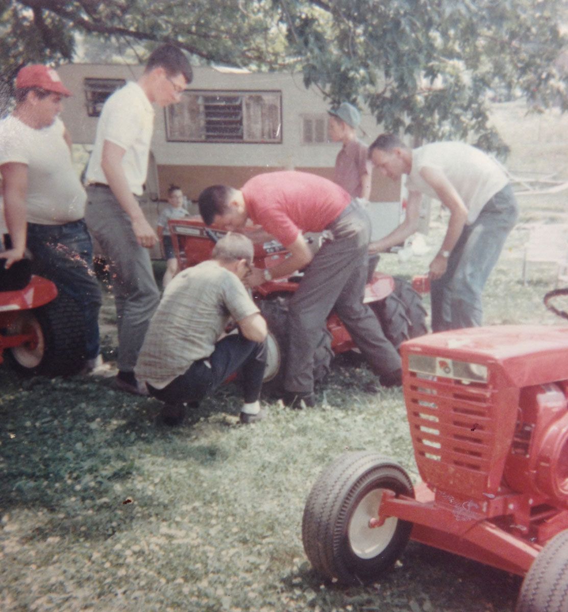 A group of men working on red tractors in a grassy outdoor setting, likely repairing them.