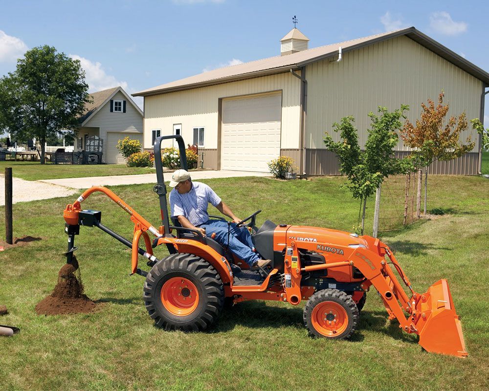 Man operates a Kubota tractor with a post-hole digger, drilling into the ground outdoors near a building.