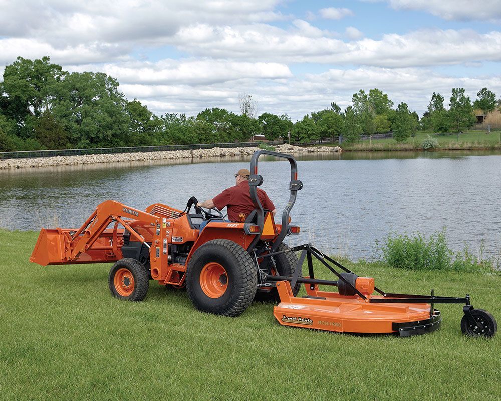 Orange tractor with operator mowing grass near a body of water.