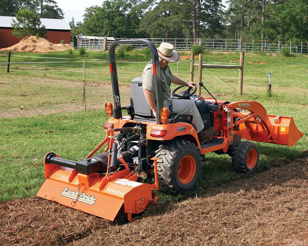 Man operating a small orange tractor with a tiller in a field.