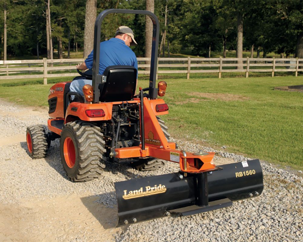 Person operating an orange tractor with a Land Pride grader on a gravel driveway near a fence.