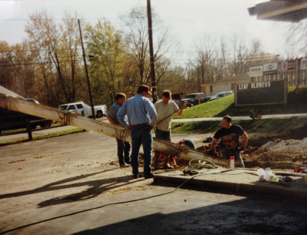 Men installing a large signpost in a parking lot. Brown sign, gray pavement, fall trees.